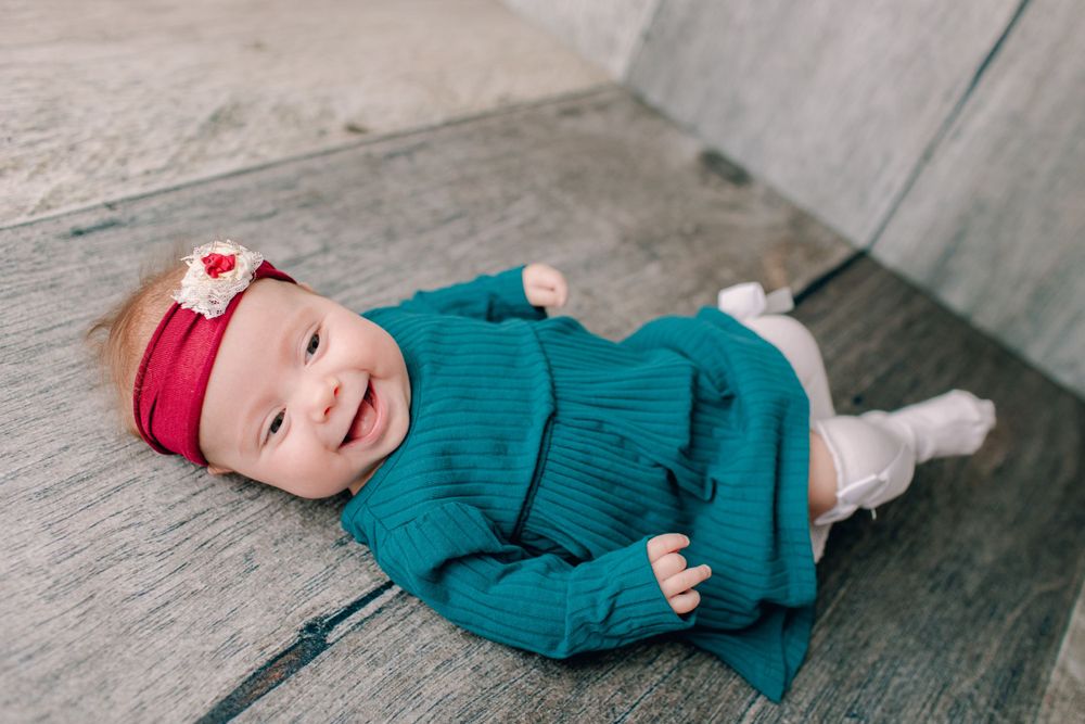 Happy baby in teal outfit and red headband lying on wooden surface.