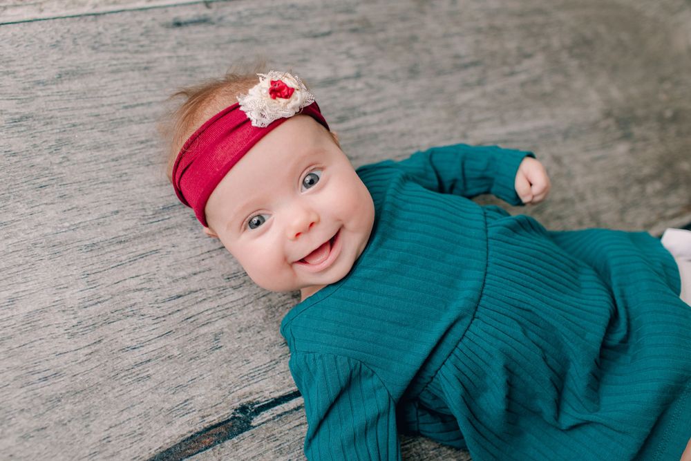 Baby in teal dress with red headband smiling on wooden background.