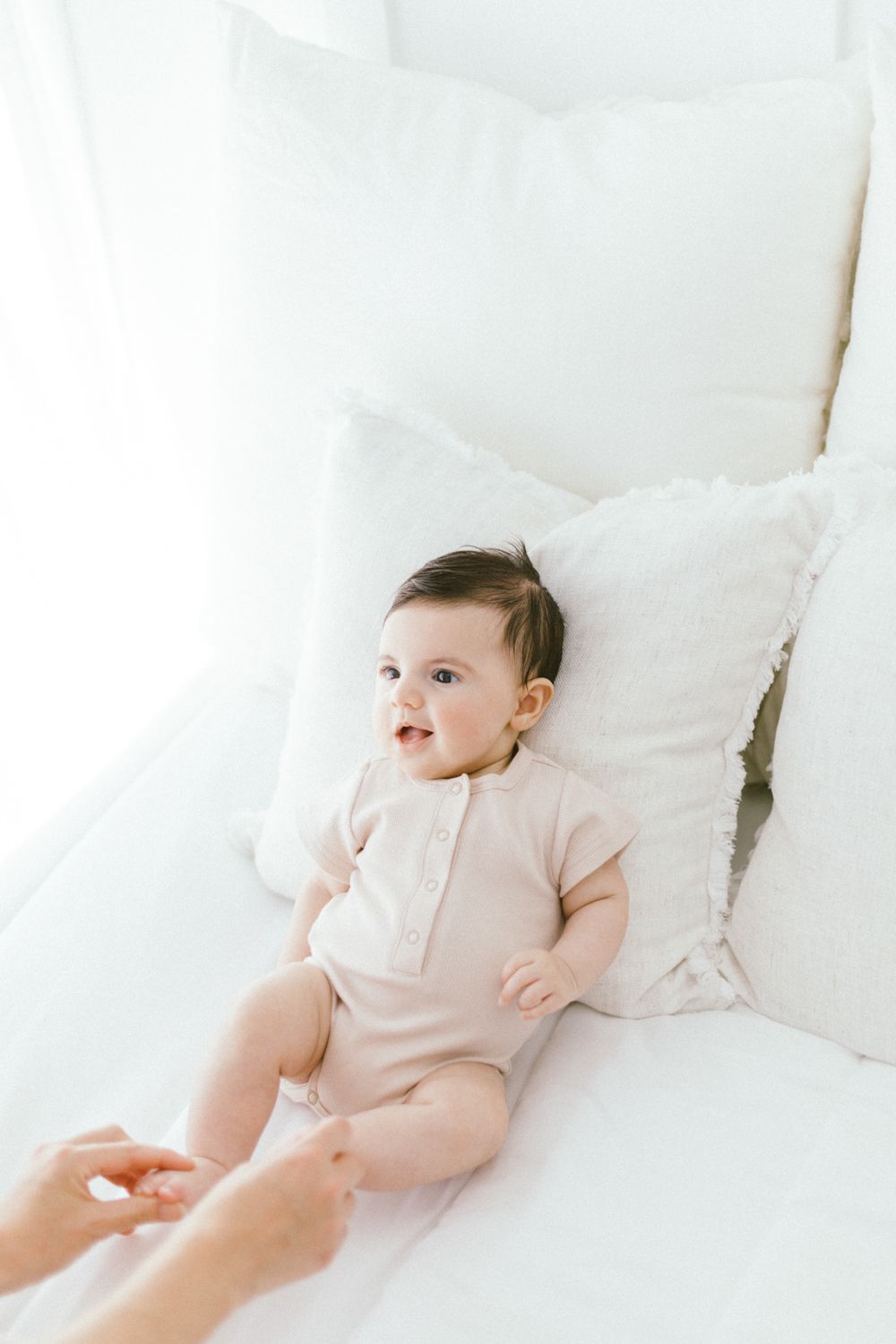 Baby in pink onesie lies among plush white pillows and bedding.
