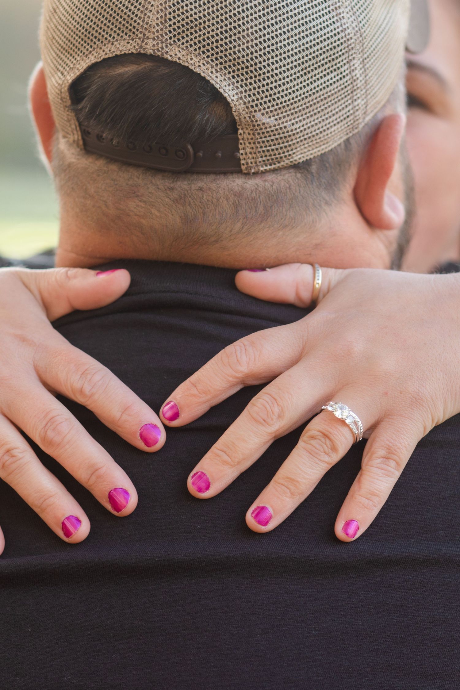 Hands with bright pink nail polish and an engagement ring rest on a black shirt.