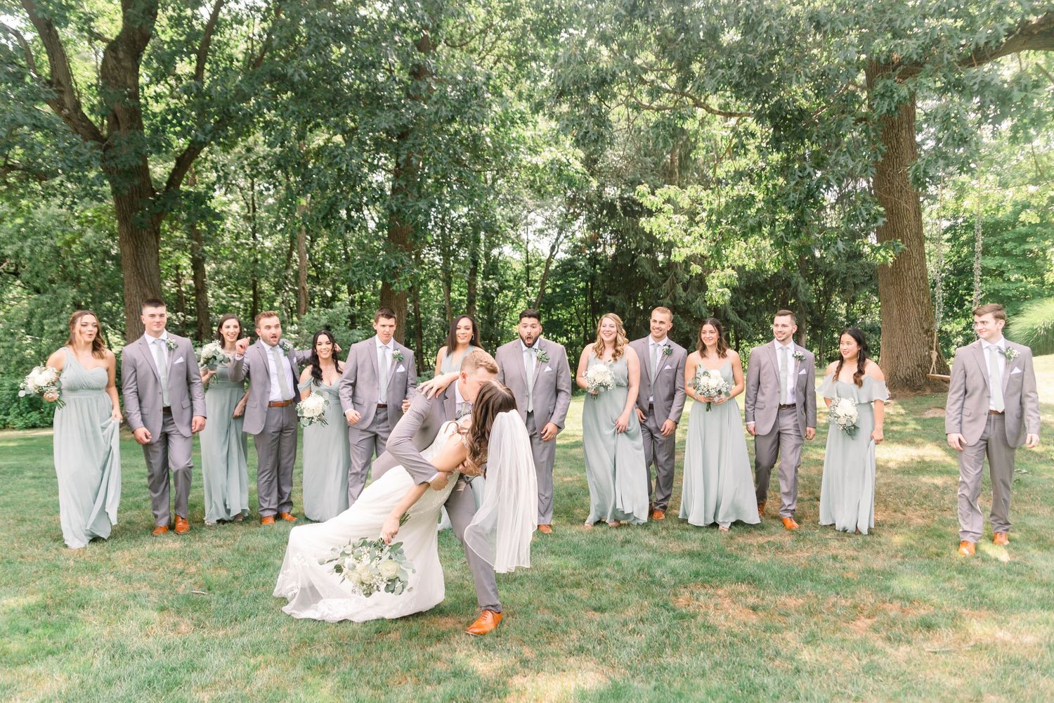 Large wedding party in gray and white attire poses for group photos in a sunny outdoor garden setting.