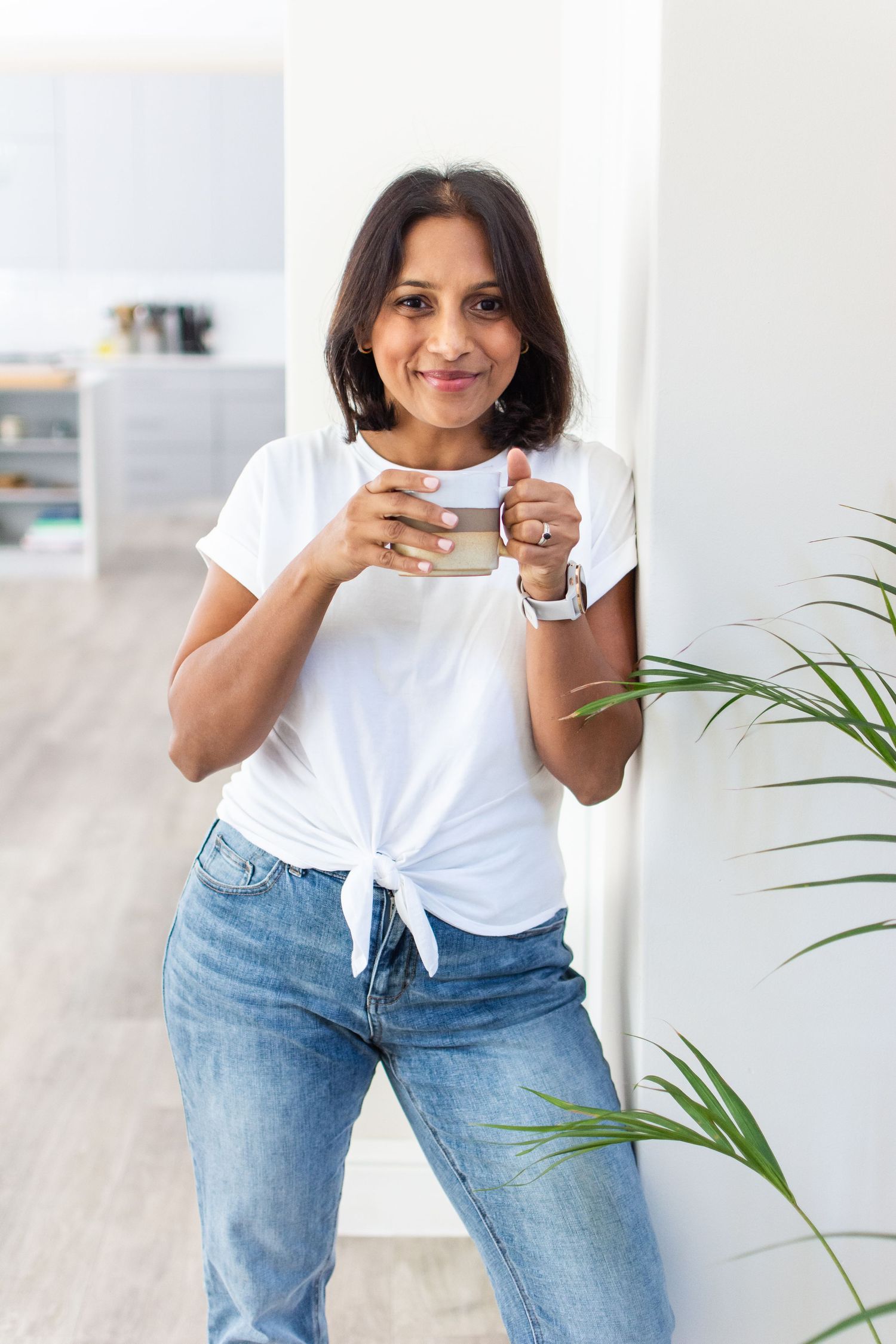 Casual lifestyle portrait holding coffee cup in white tied t-shirt and jeans near indoor plants.