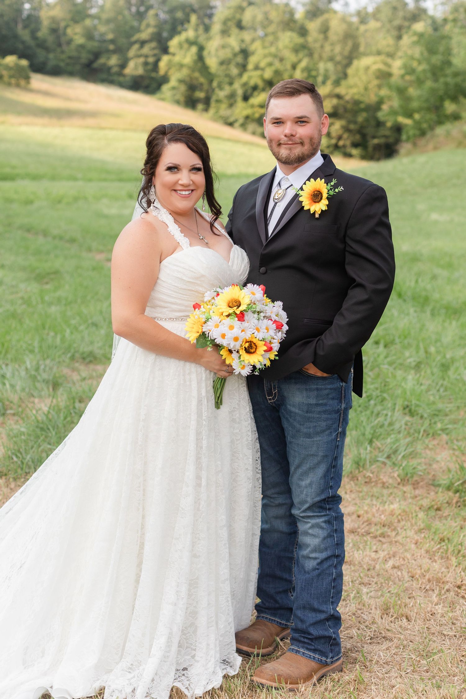 Rustic outdoor wedding portrait with sunflower bouquet against rolling hills landscape.