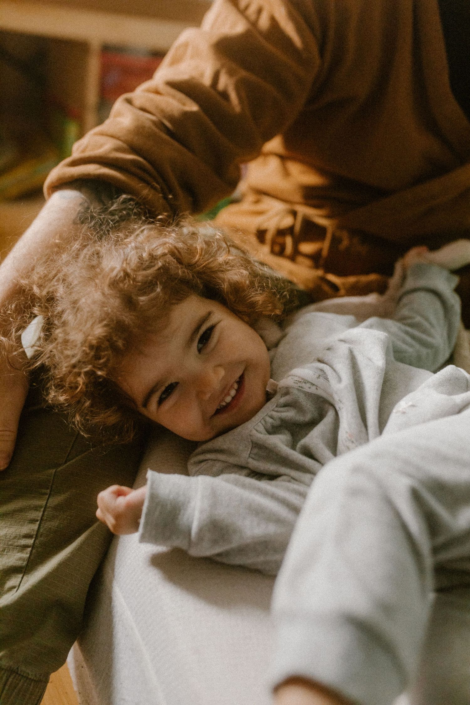 Candid shot of toddler with curly hair lying down and smiling warmly in natural lighting.