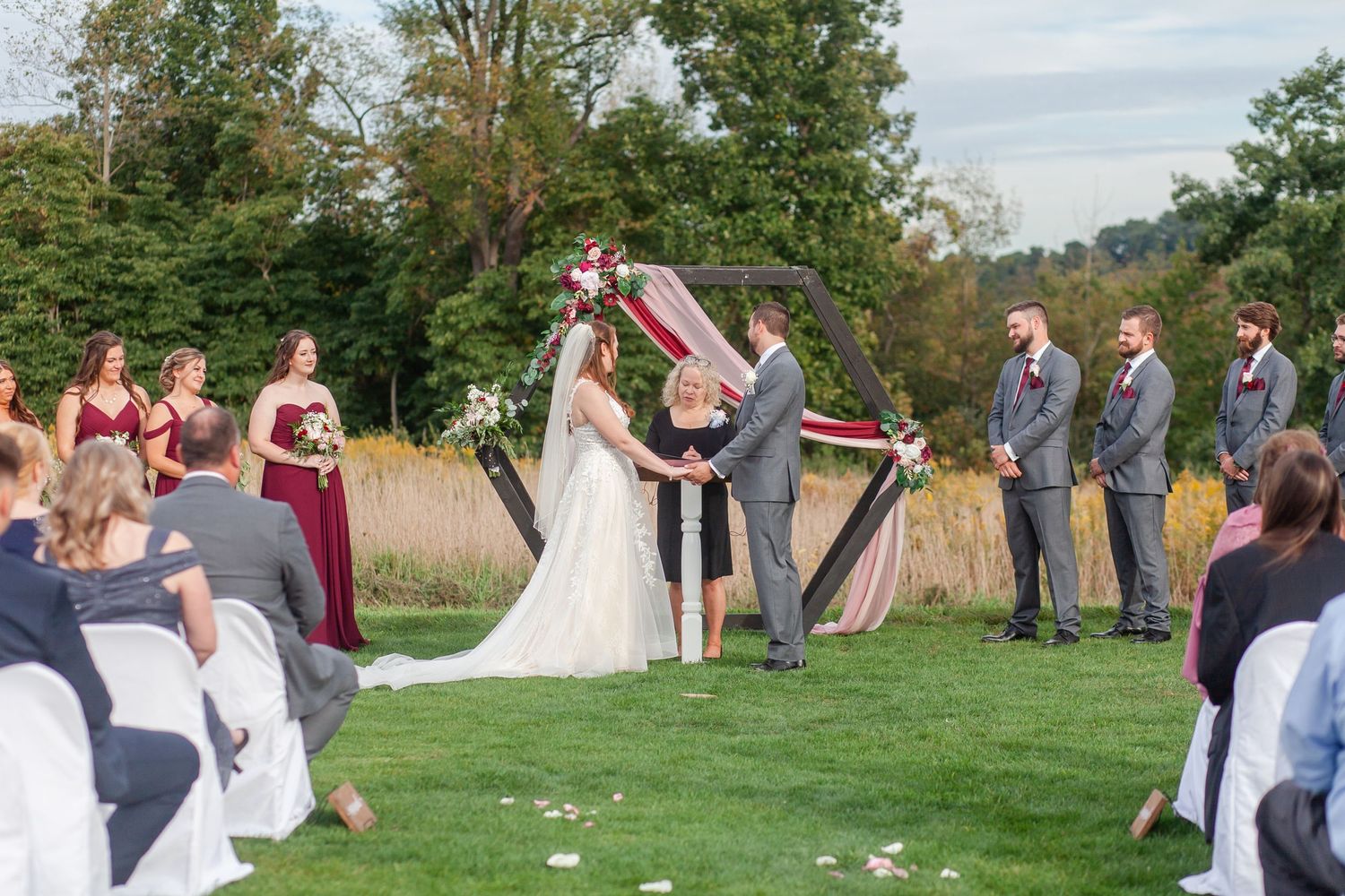 Wedding guests seated on white chairs watch an outdoor ceremony with burgundy and white decor elements.