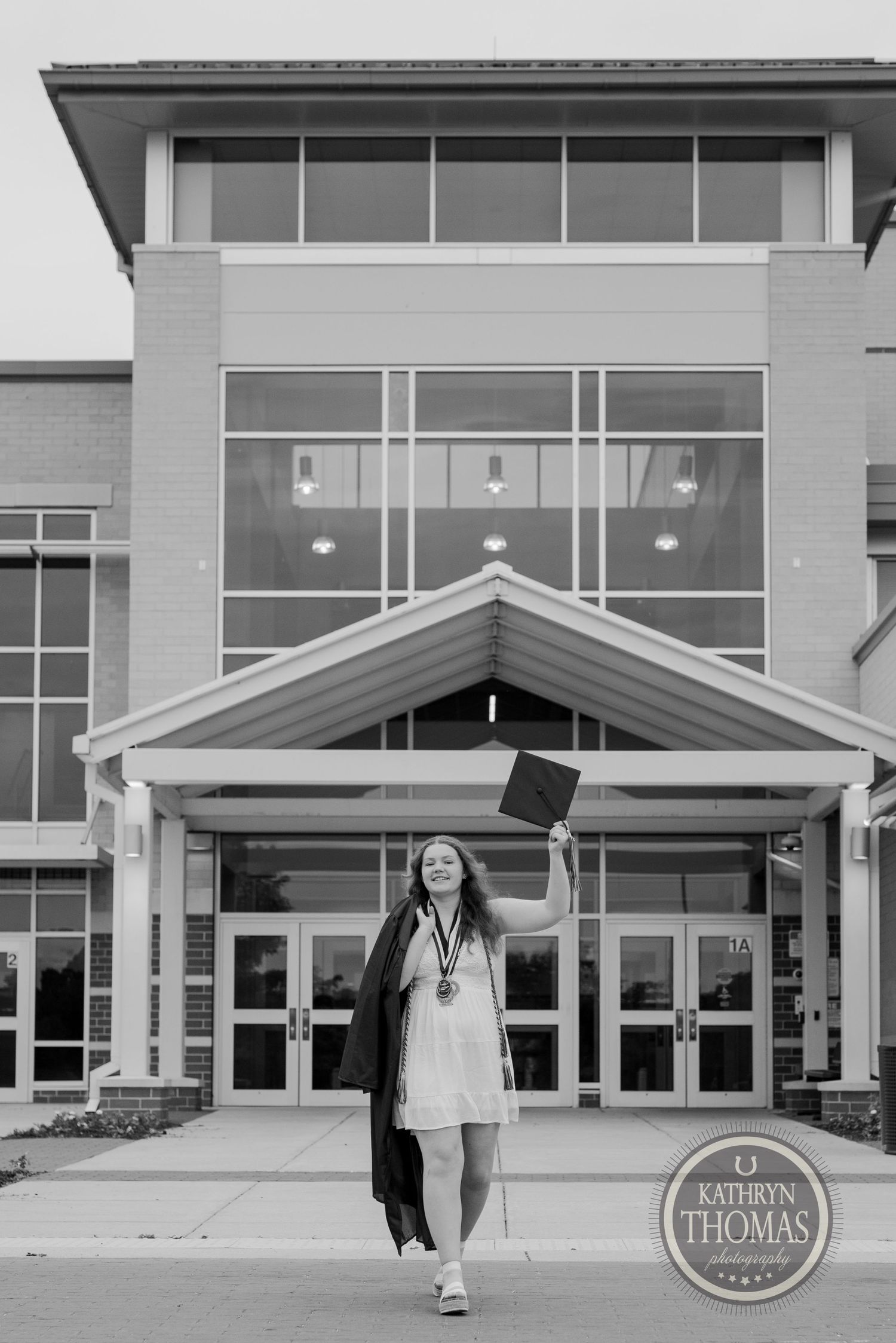 Black and white photo of graduate jumping with cap outside modern school entrance.