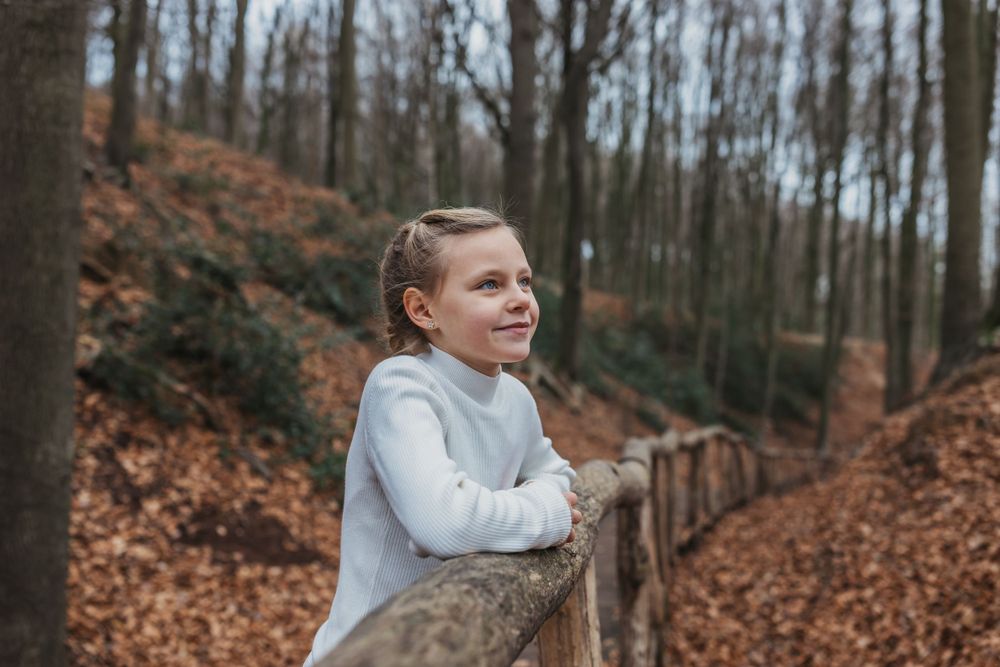 Person in white sweater smiles while leaning on wooden railing in autumn forest setting.
