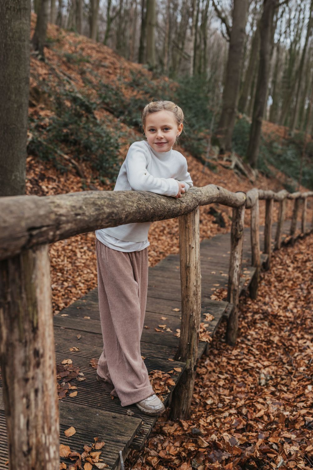 Person in white sweater and pink pants stands on wooden forest walkway during autumn.