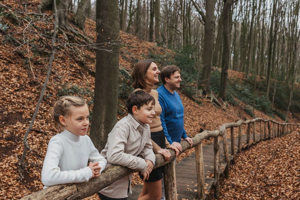 People lean against a rustic wooden railing on an elevated forest path covered in fallen leaves.