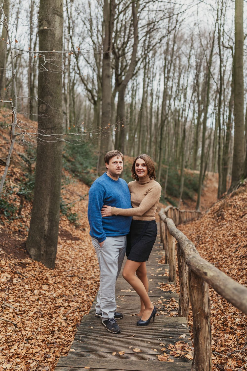 A couple embraces on a wooden walkway surrounded by fall foliage in the forest.