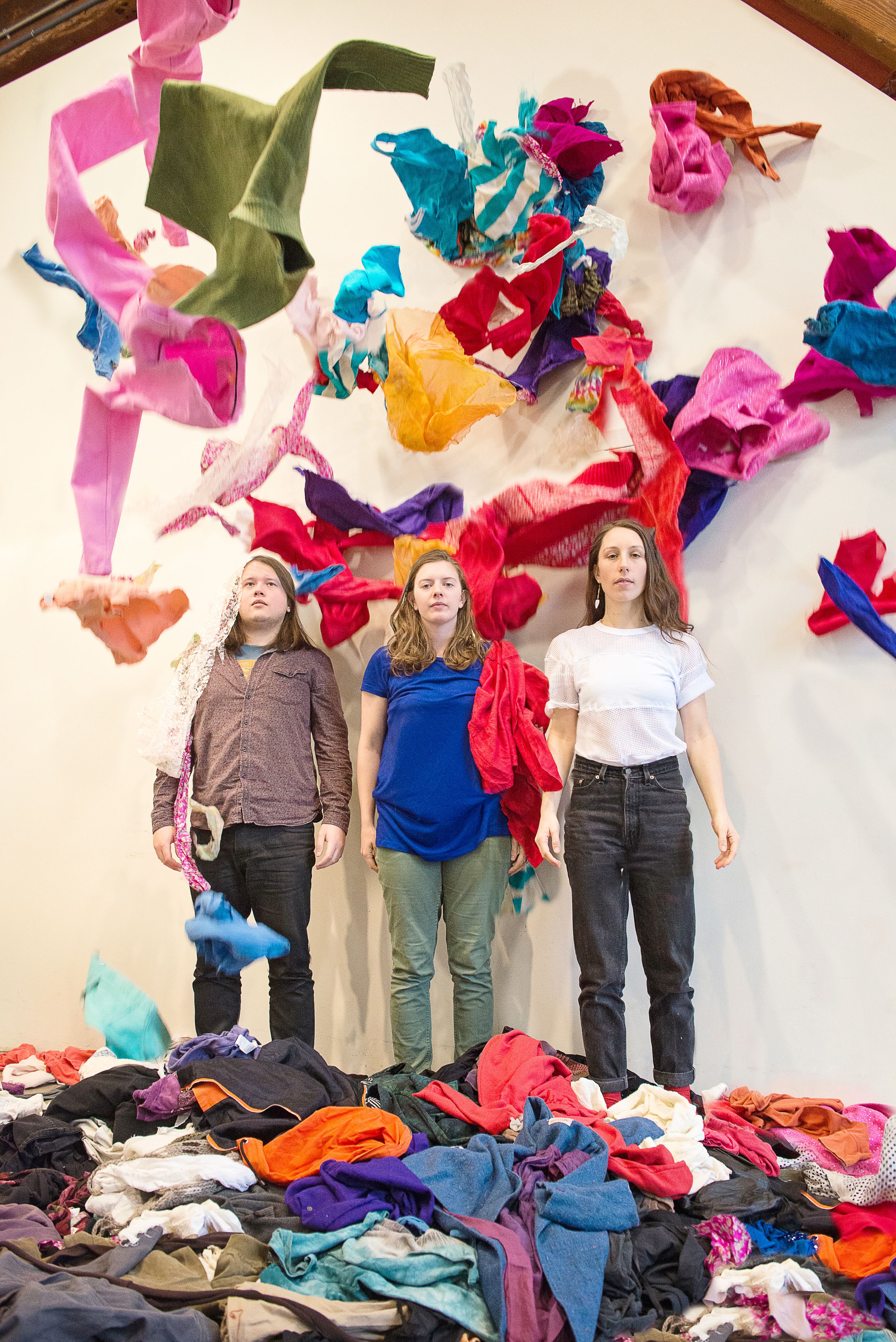 Three people stand against a wall with colorful fabric pieces floating above them. Photography by Riz Herbosa