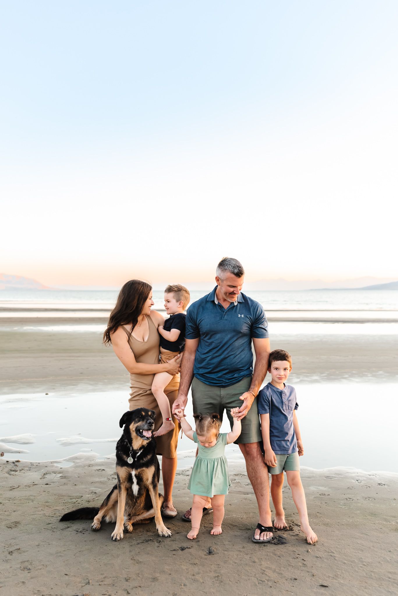Family and their German Shepherd dog pose playfully together on a scenic beach at sunset.