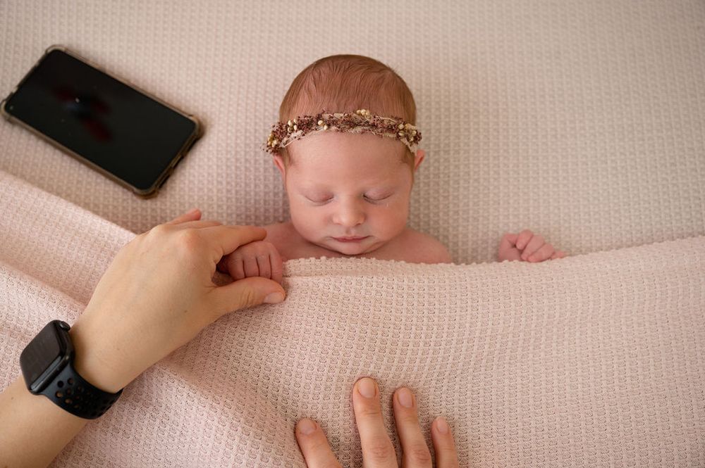 Sleeping newborn with delicate headband on cream blanket with hands and phone visible.