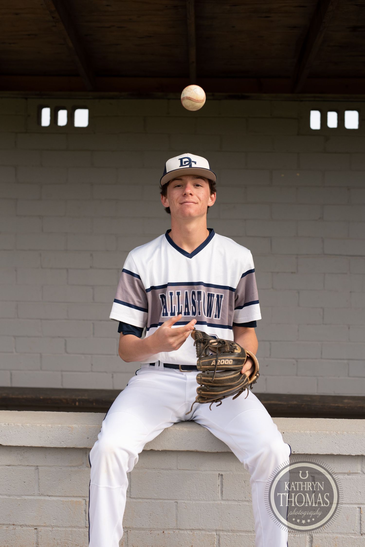 Baseball player tosses ball while sitting in dugout wearing Dellasoft uniform.