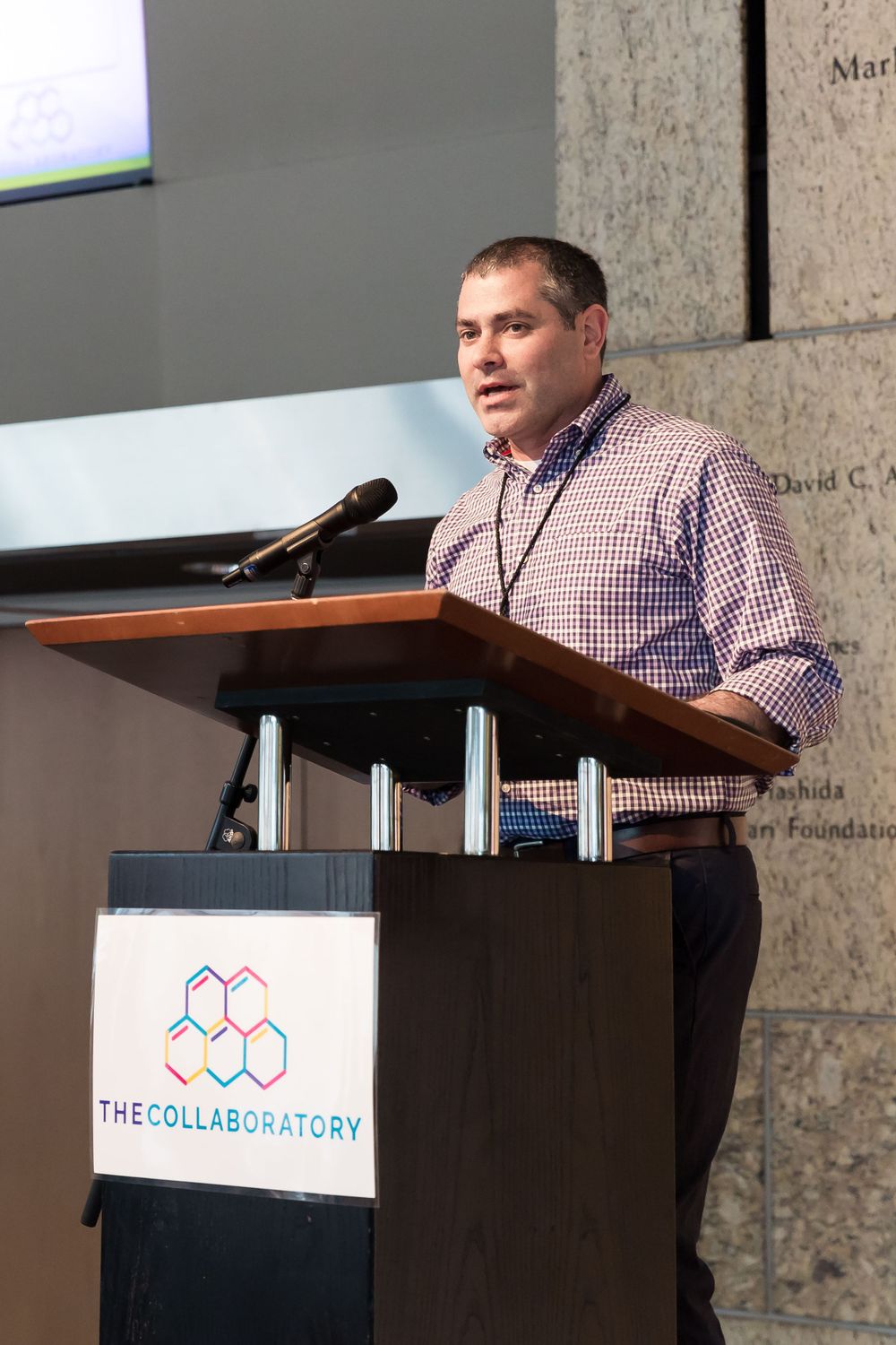 A speaker presents at a podium with The Collaboratory logo displayed. Mika Larson Photography.