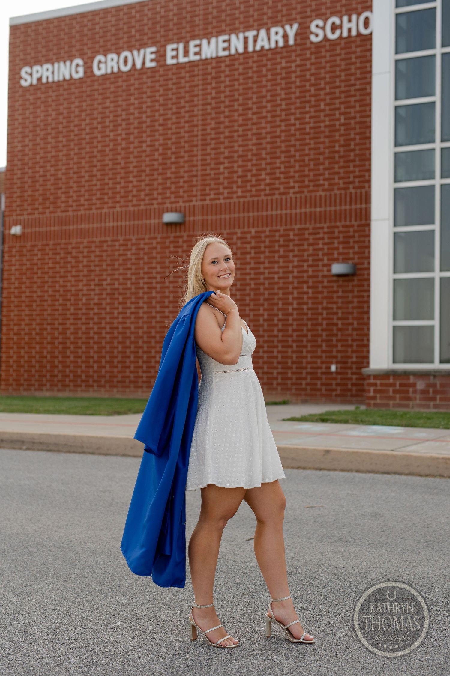 Graduate in white dress and blue gown poses outside Spring Grove Elementary School at sunset.