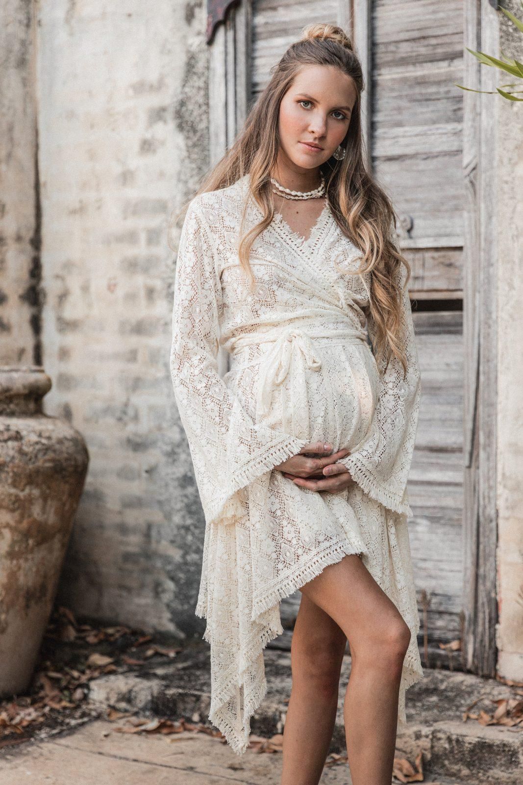 Elegant maternity portrait featuring woman in white lace dress against rustic backdrop.