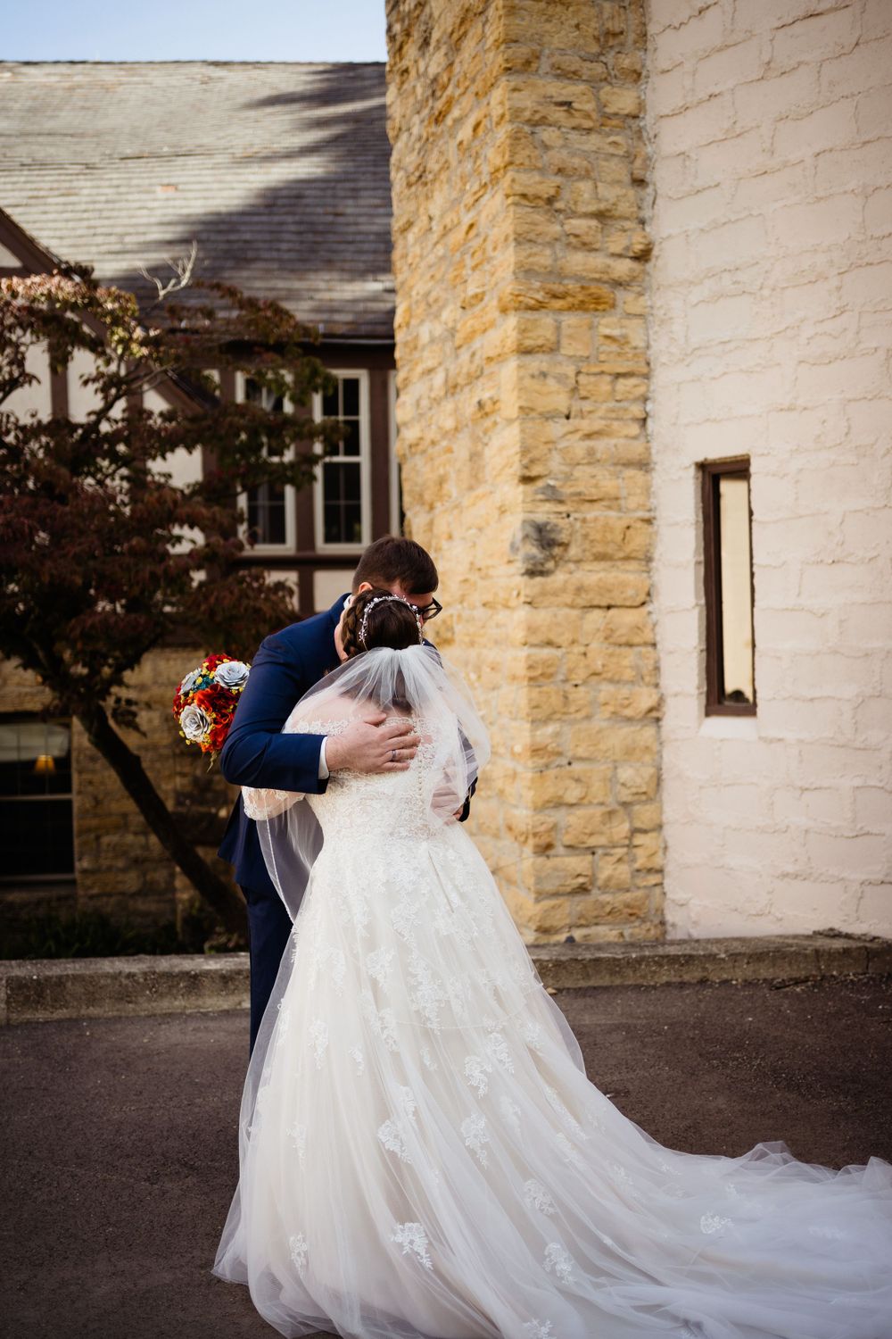 A couple embraces outside a stone building on their wedding day, with the bride wearing a flowing white lace gown.