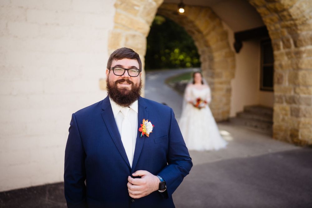 A groom in navy suit and glasses stands under stone archway at wedding venue.