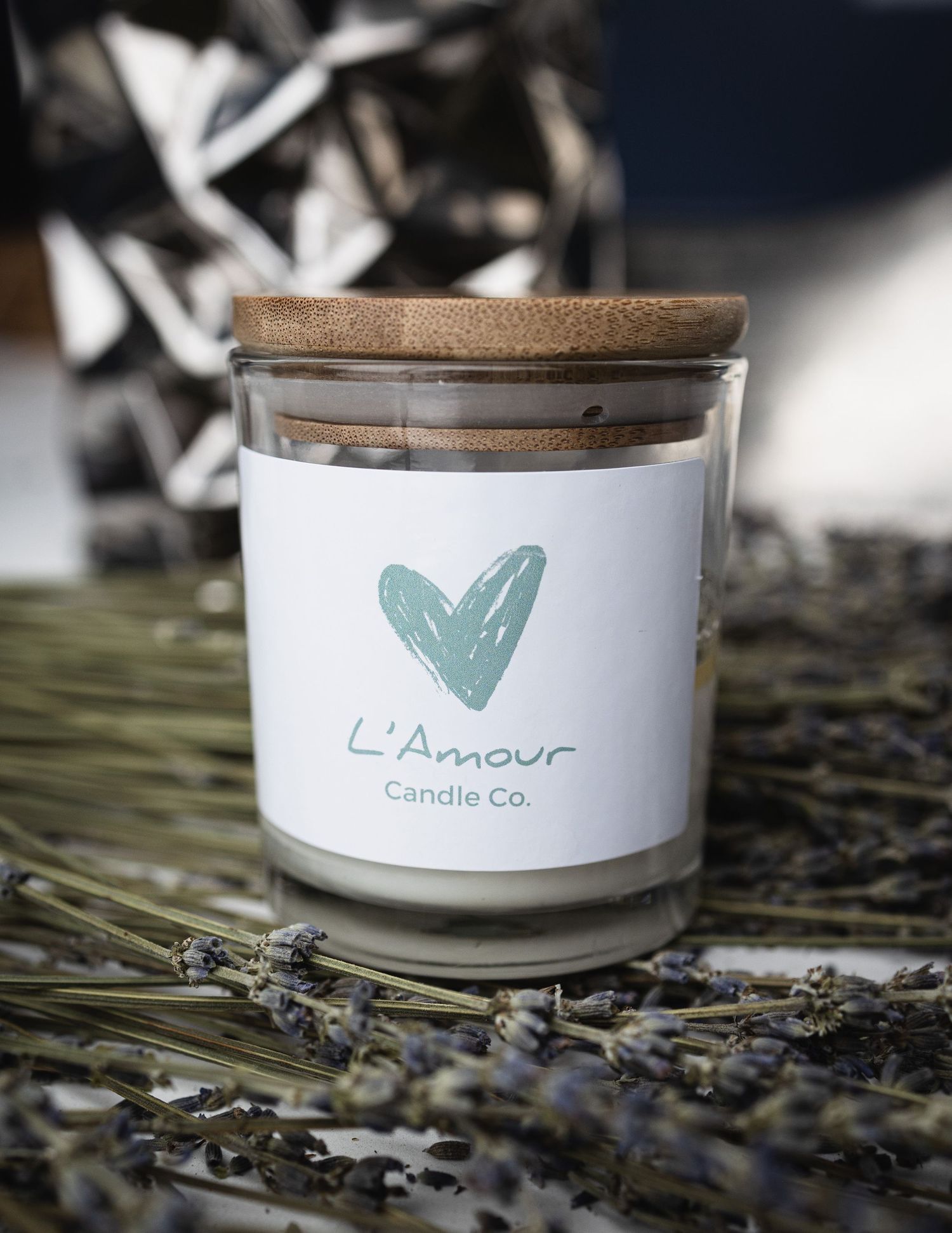 A white candle jar labeled L'Amour with a teal heart logo surrounded by dried lavender sprigs.