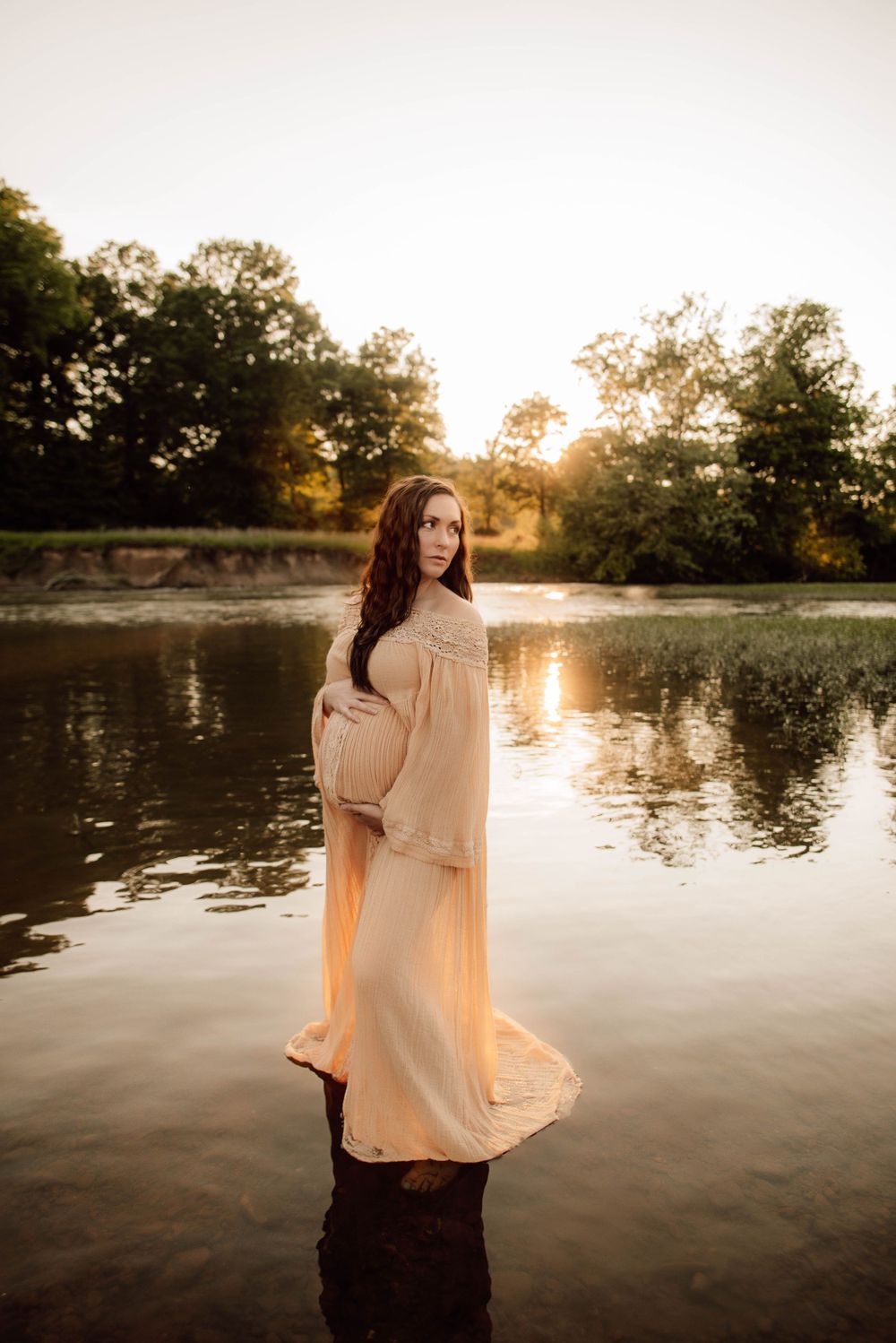 Woman in flowing dress stands by tranquil lake at golden hour.