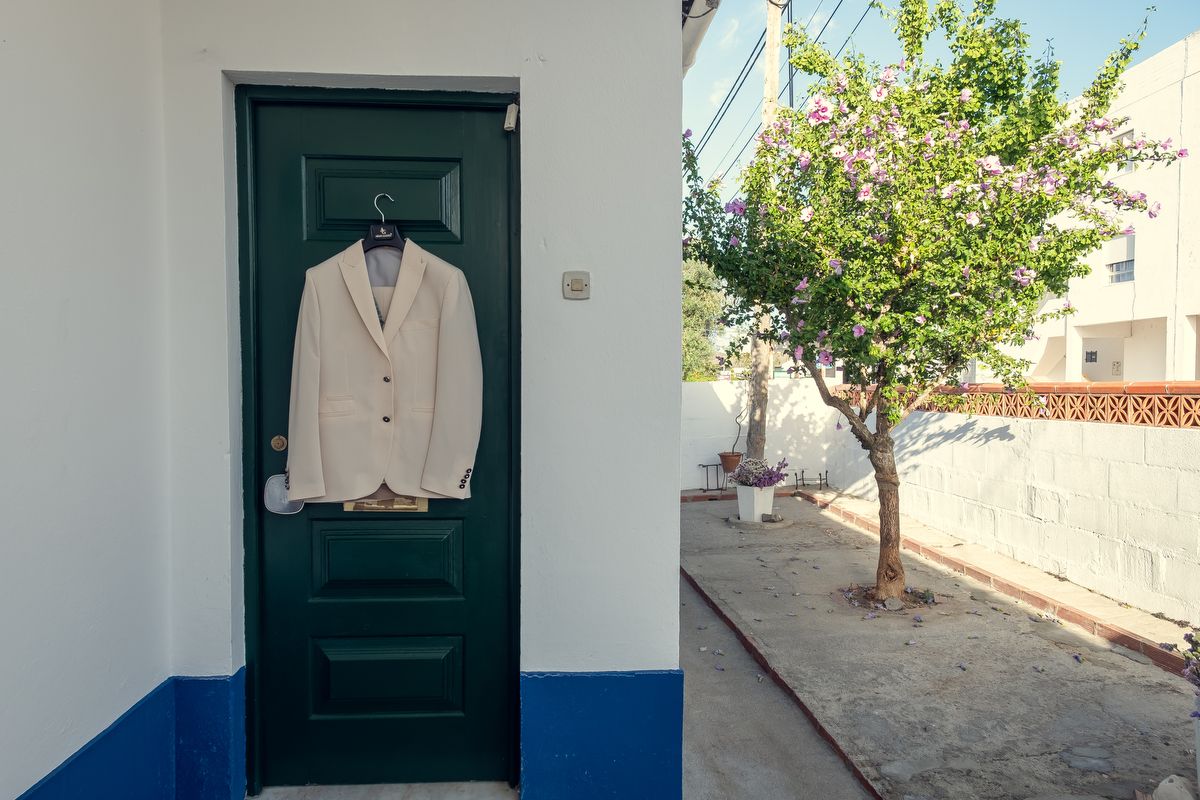 Elegant beige wedding suit jacket hanging on a green door outside a white Mediterranean house.