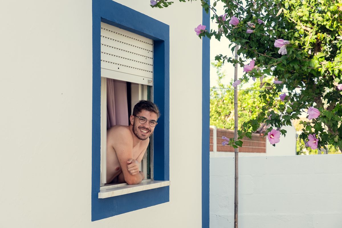 Smiling shirtless man leans out of a blue-framed window surrounded by blooming pink flowers.