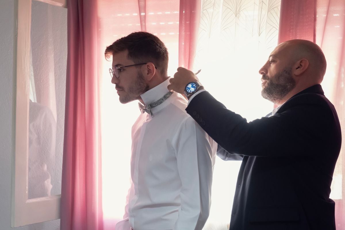 Older man adjusts the collar of a young groom dressed in white near a pink curtained window.