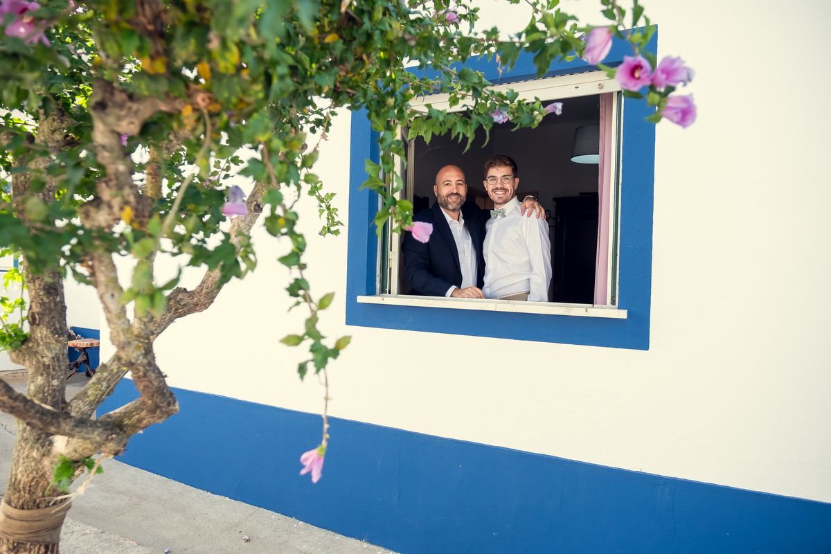 Two smiling men pose together at a blue-framed window beside a flowering tree at a wedding.