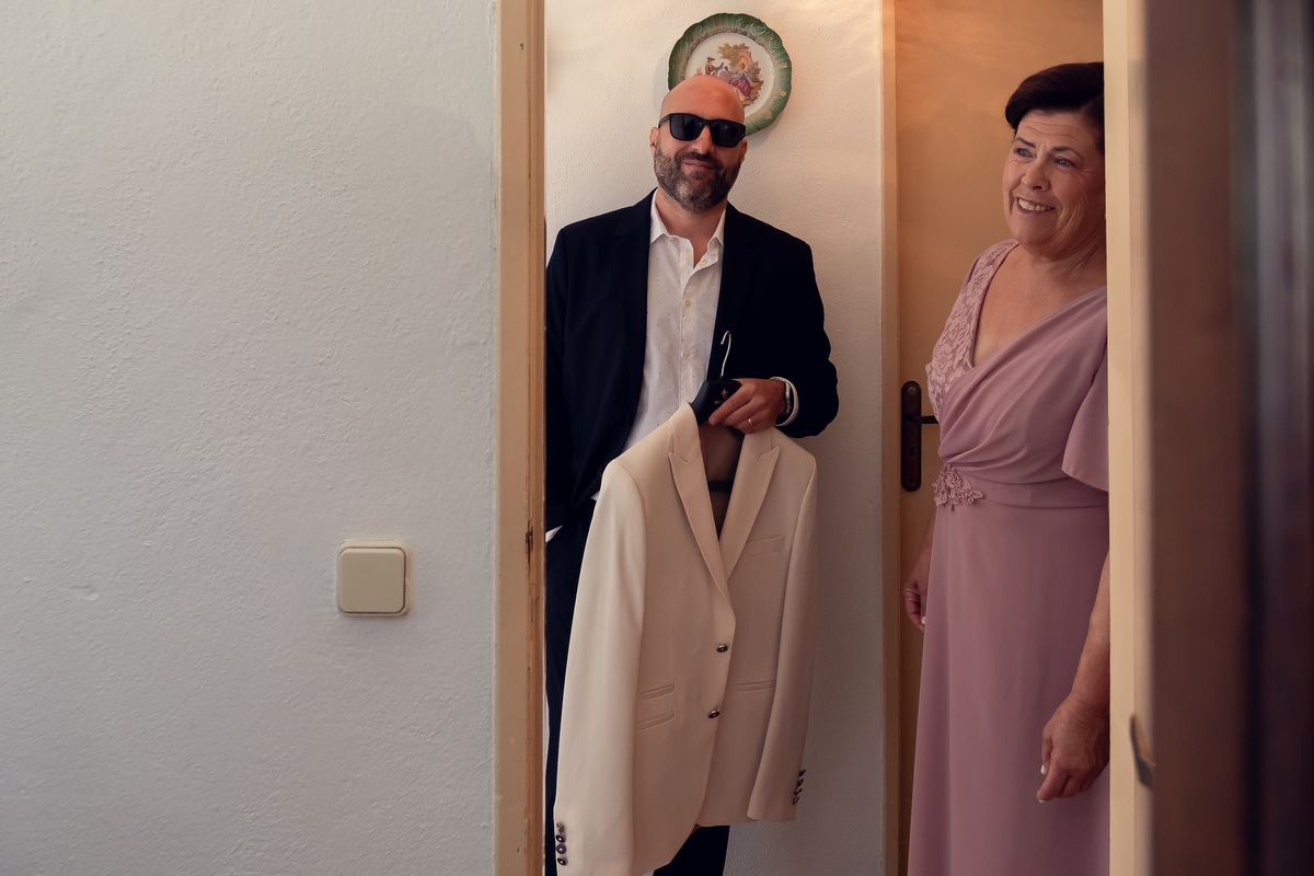 Man in sunglasses holding white blazer smiles at woman in pink dress standing in doorway.