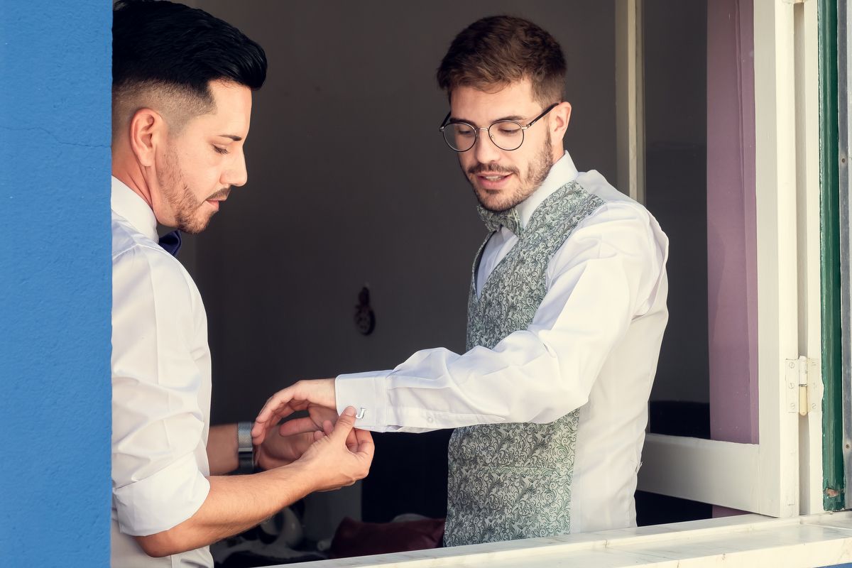 Two grooms helping each other dress on wedding day, one adjusting the other's cufflinks near a bright window.