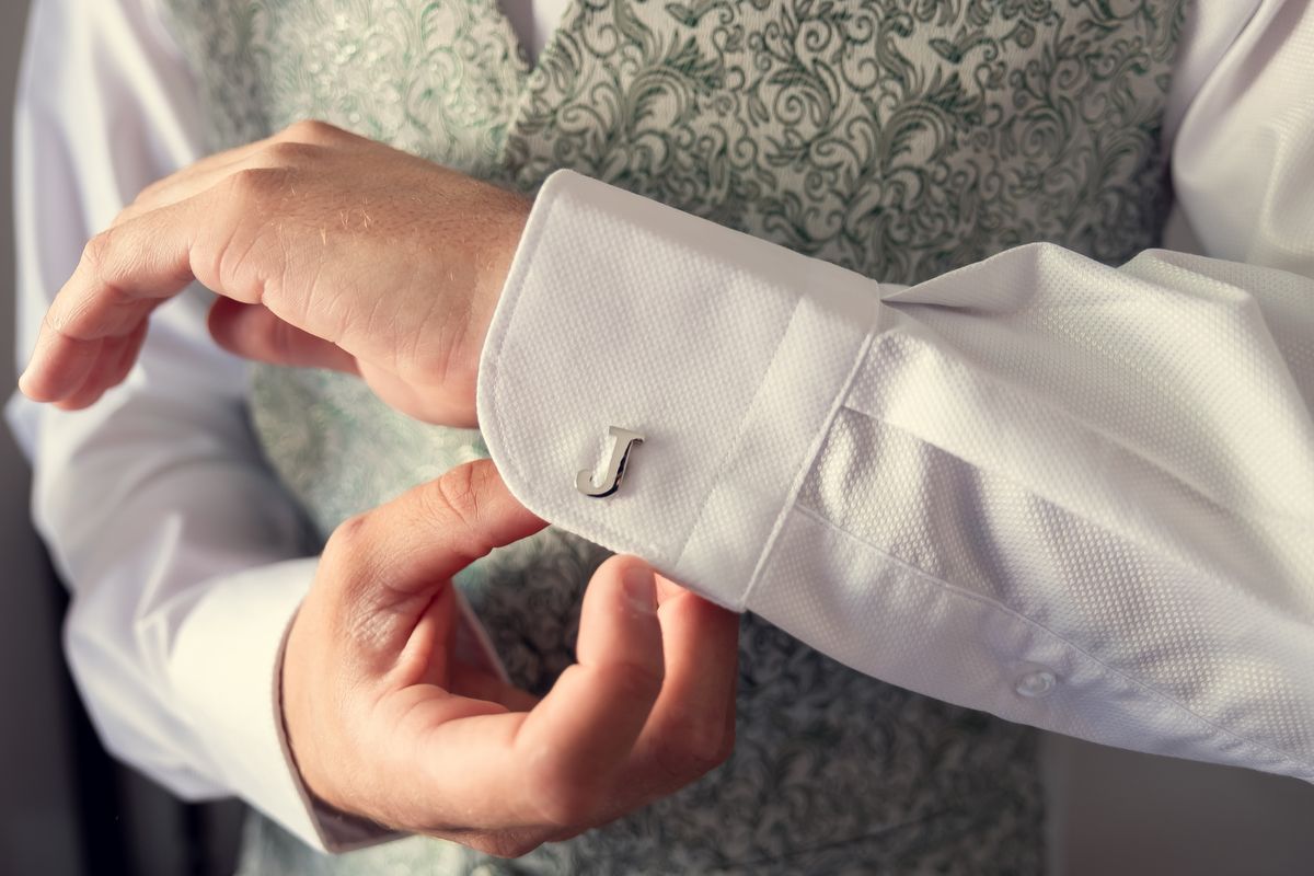 Man fastening silver cufflinks on white dress shirt with green floral vest, elegant wedding attire detail.