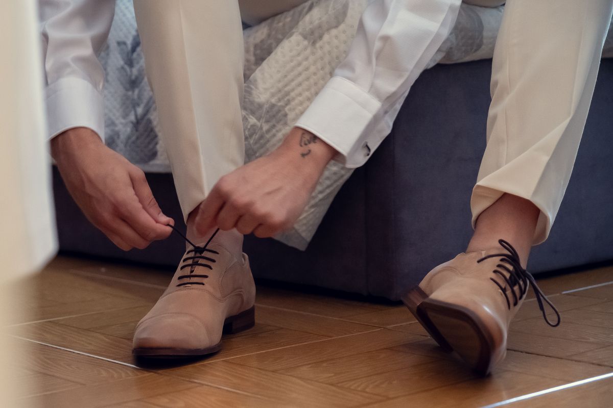 Man in white dress shirt tying brown suede oxford shoes on hardwood floor, preparing for formal event.