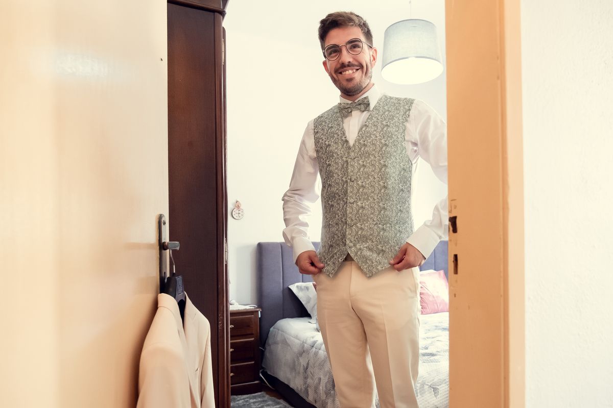 Smiling man in glasses wearing a floral vest and white shirt stands in a doorway, dressed for a formal occasion.