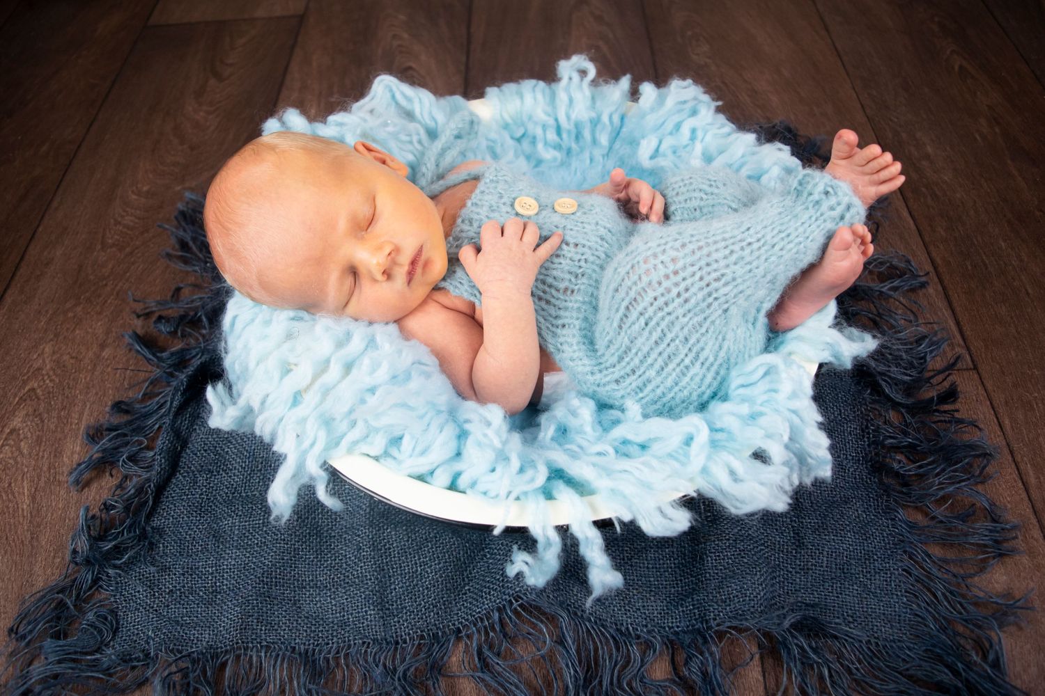 Newborn baby boy photographed in a bowl wearing a blue fluffy outfit on wooden floor taken by Heather Katie Photography.