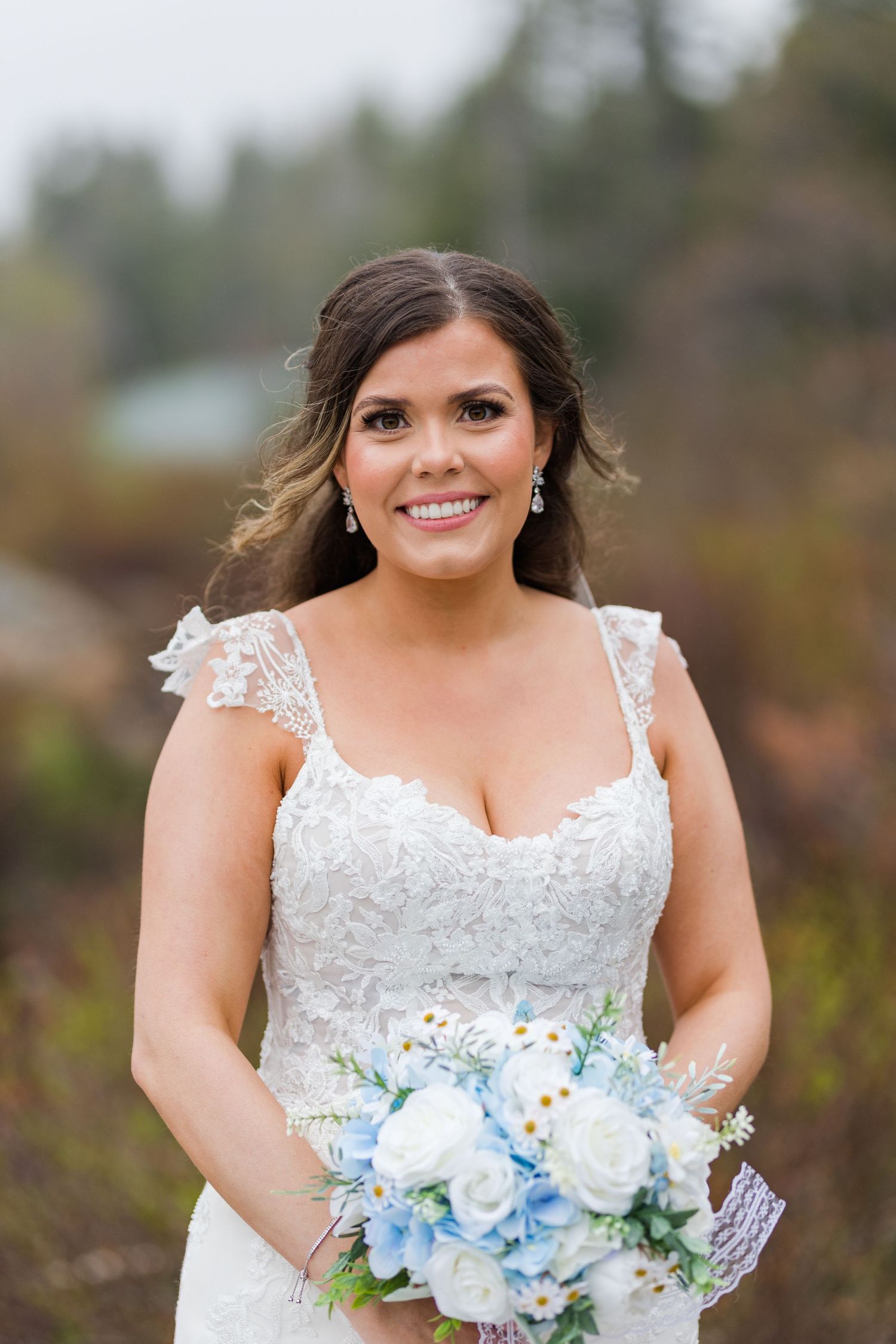 Bride in lace dress holding light blue bouquet smiles against autumn mountain landscape.