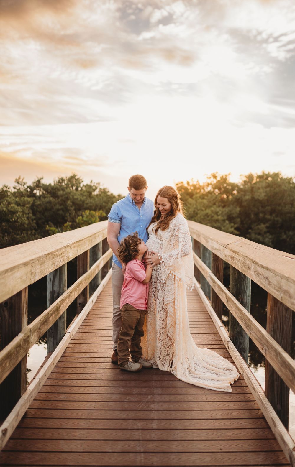 Family of three poses together on a wooden boardwalk during golden hour.