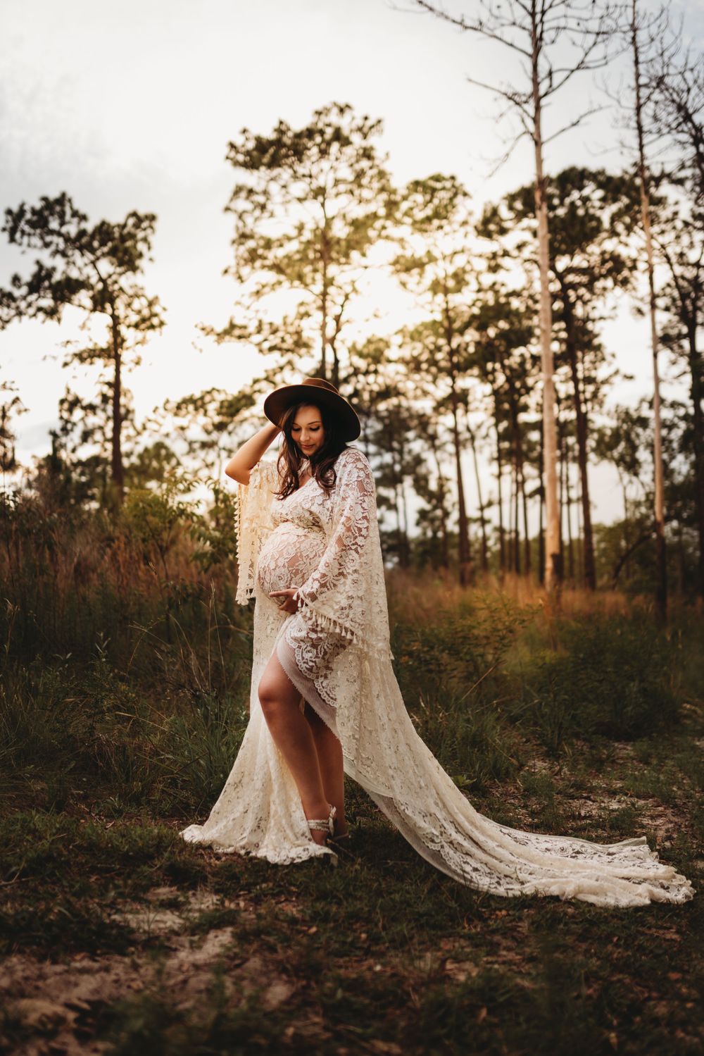 A woman in a flowing white lace dress poses in a sunlit forest.