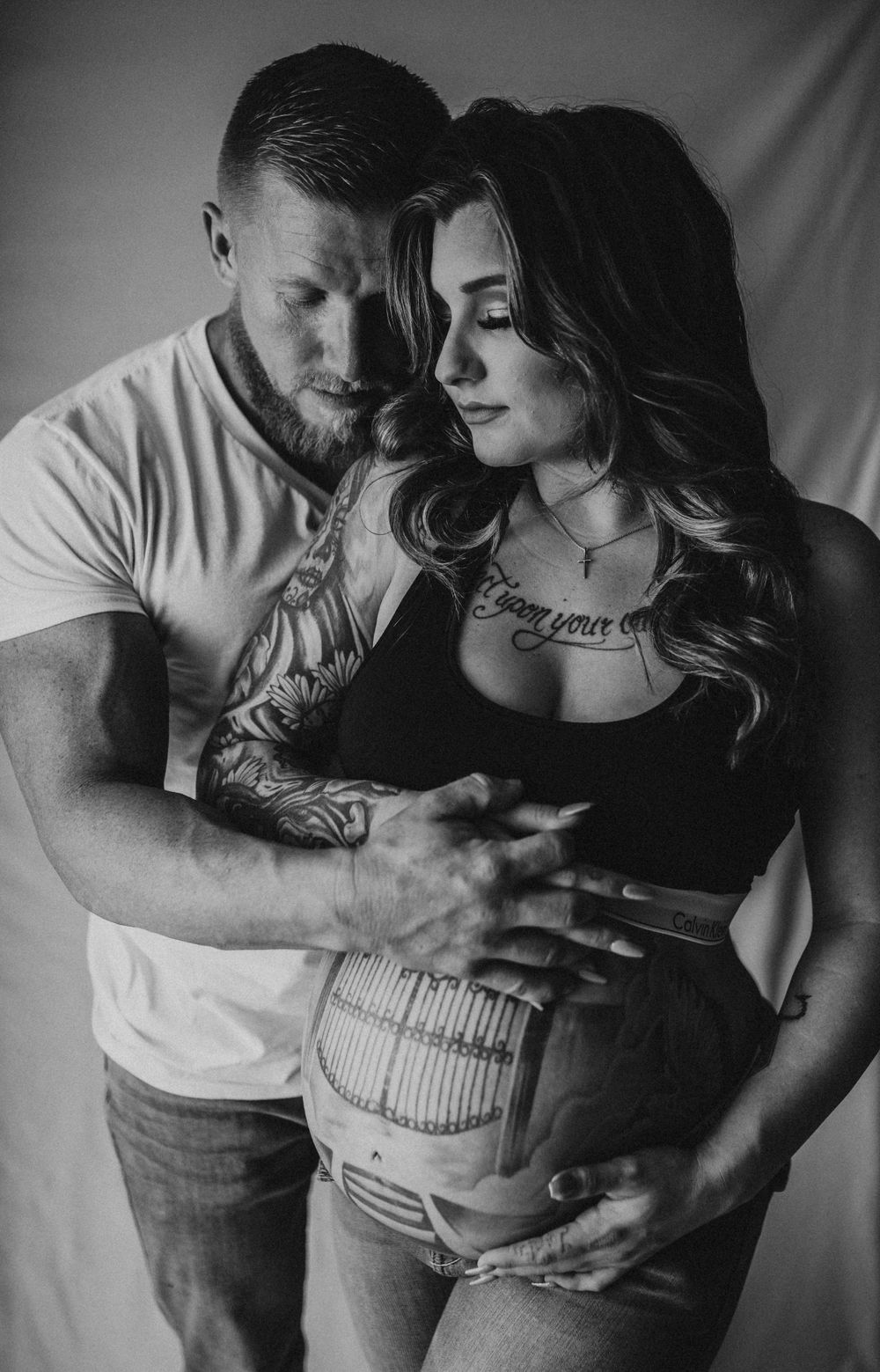 A moody black and white maternity photo showing a couple in intimate embrace with painted belly art.