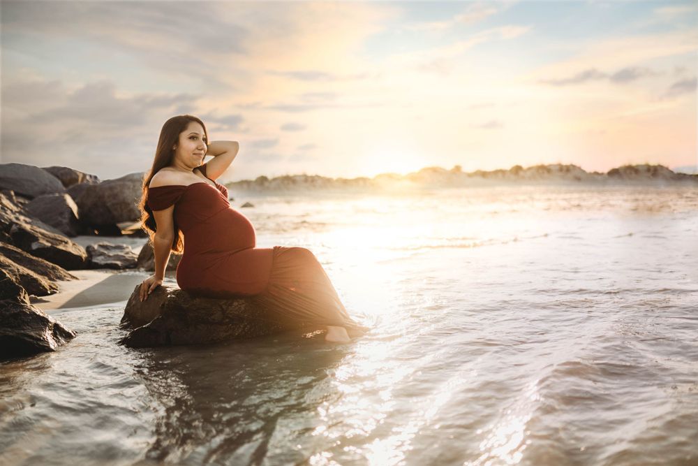 Woman in red dress sitting on coastal rocks during golden hour as waves crash nearby.
