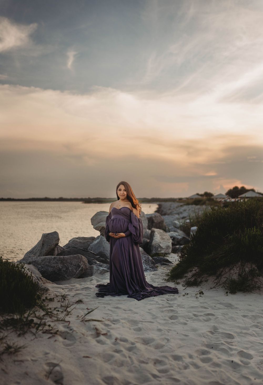 Woman in purple gown poses on beach with rocks and water during sunset.