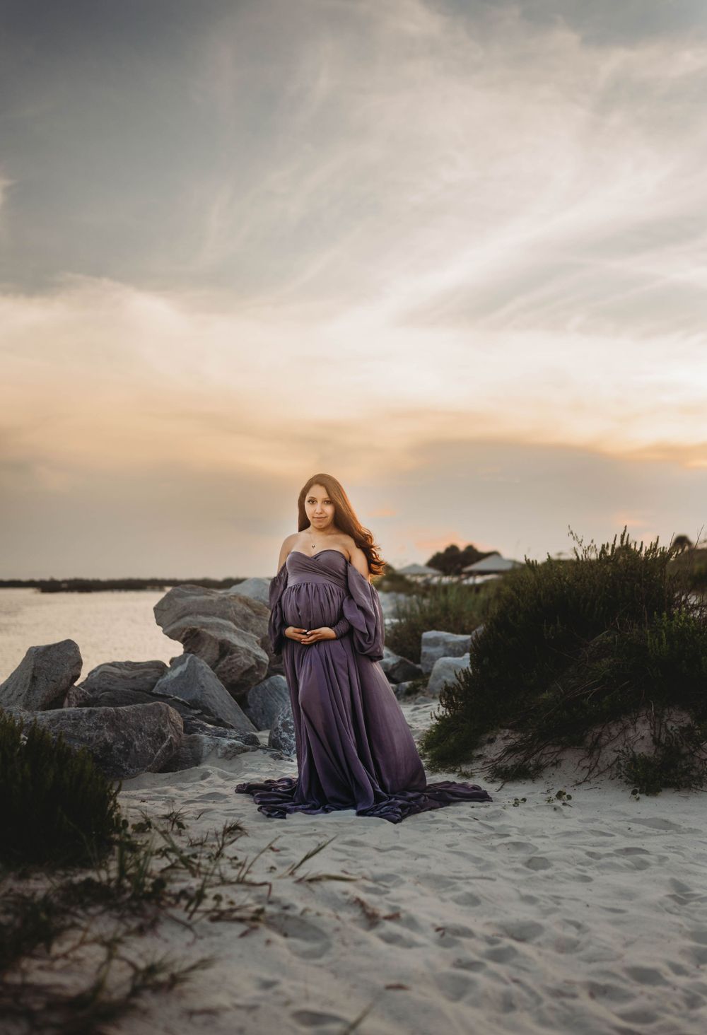Woman in flowing purple dress stands on sandy beach during dramatic sunset.