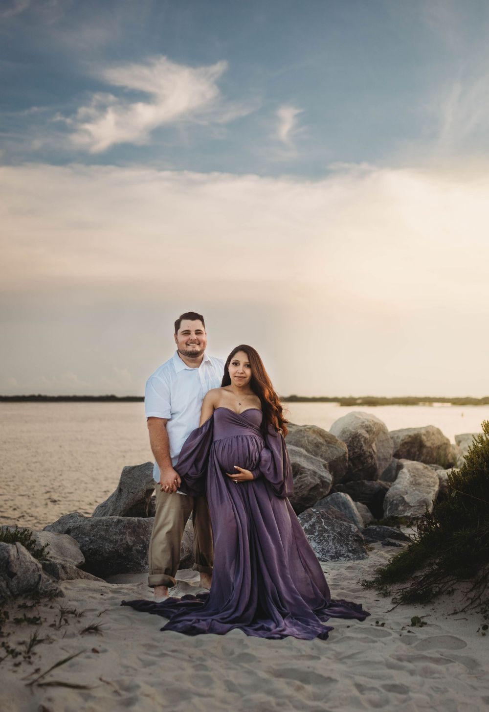 Couple stands among beach rocks during sunset maternity photoshoot.