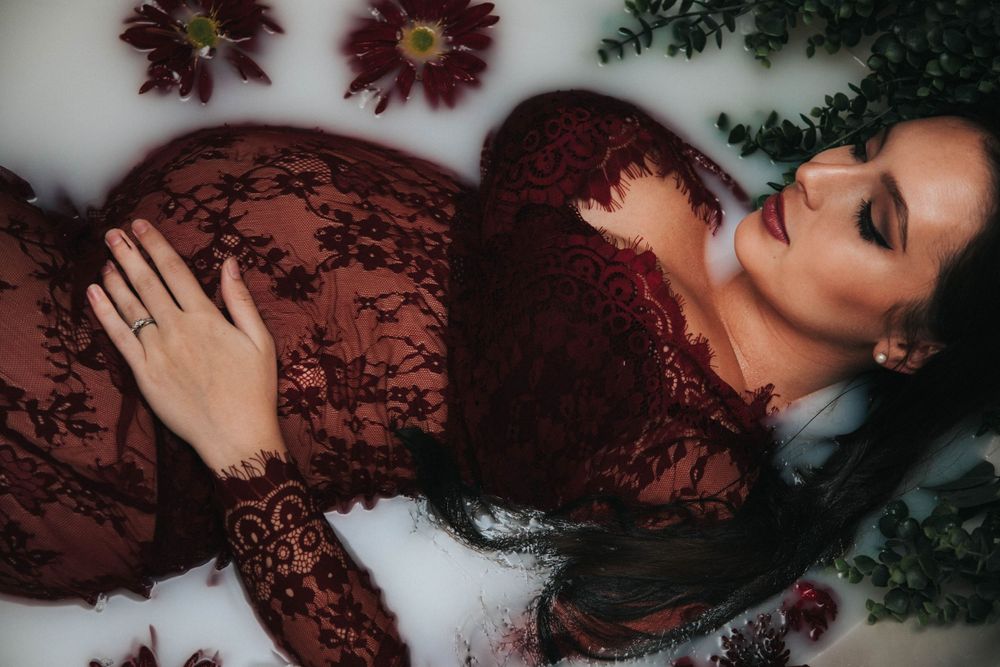 Ethereal maternity portrait with burgundy lace dress surrounded by flowers in milky bath water.