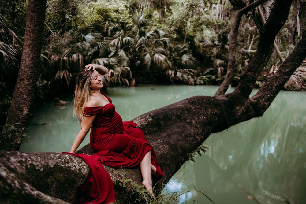 Artistic portrait in red dress sitting on tree branch over tranquil green water.