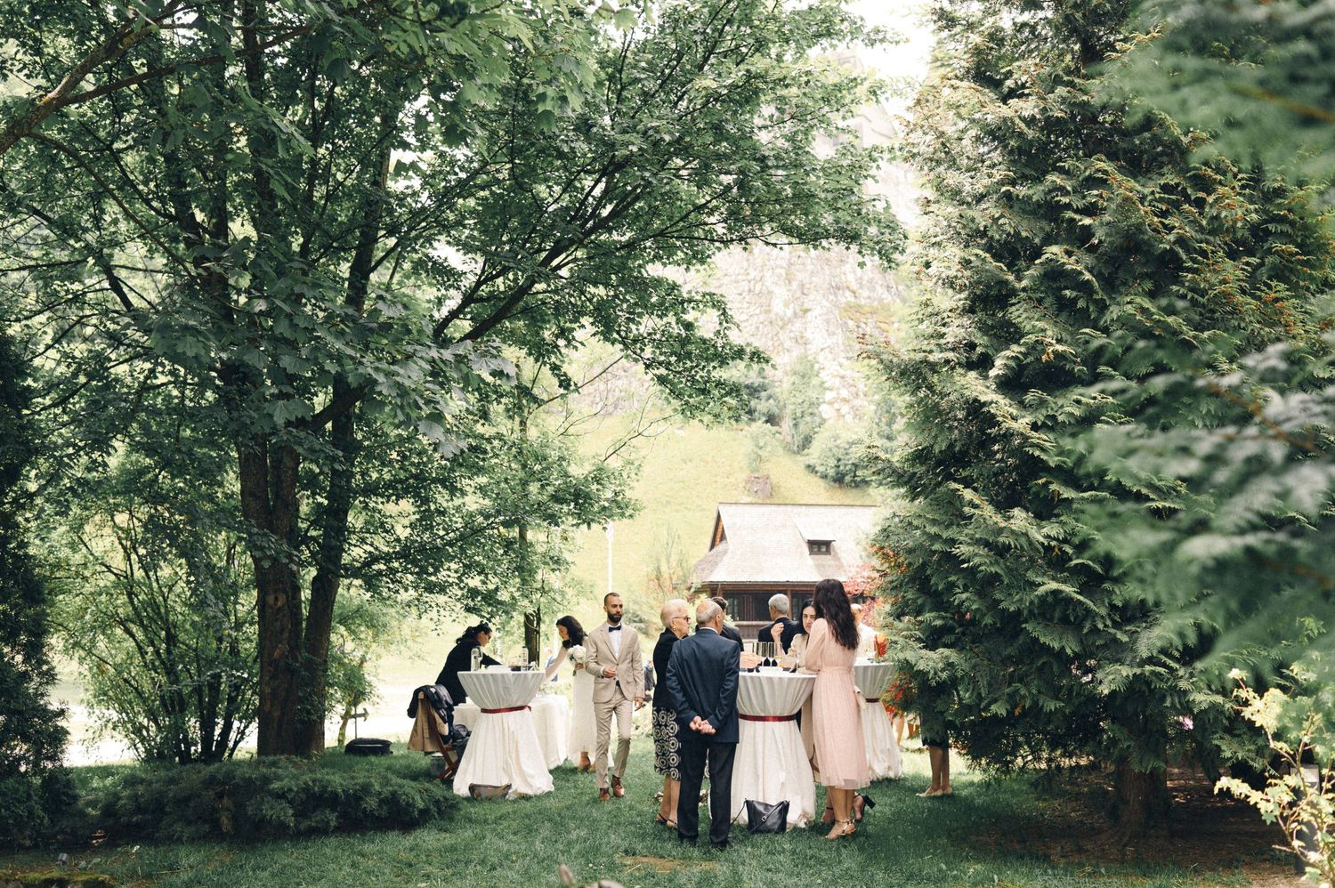 Wedding guests gather on a lush green lawn during an outdoor ceremony surrounded by trees.
