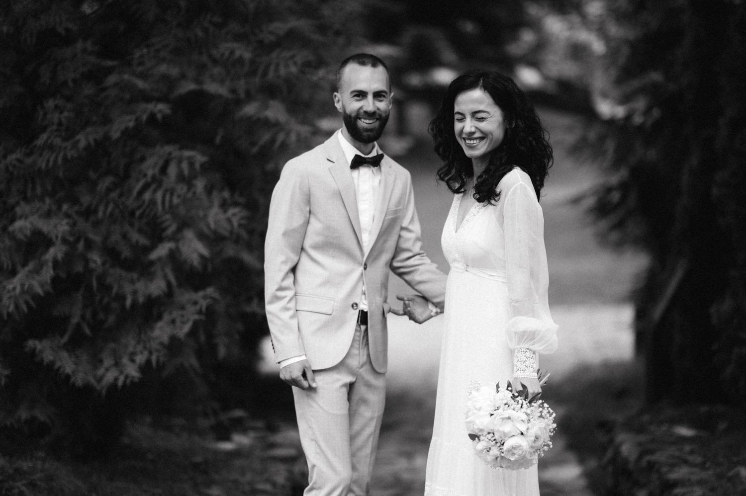 Black and white portrait of a couple in wedding attire standing among evergreen trees.
