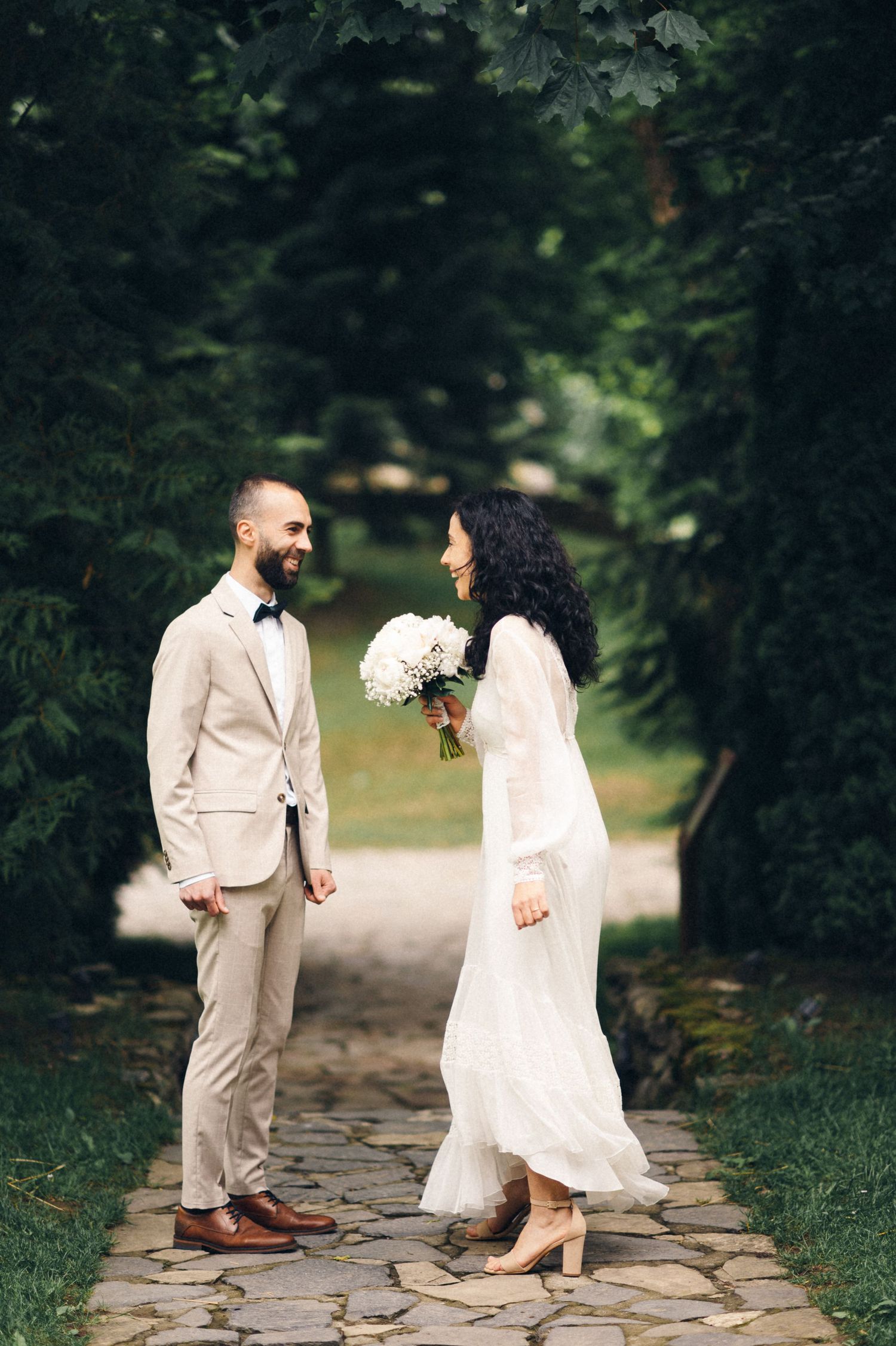 A couple in wedding attire standing on a stone path surrounded by green foliage.