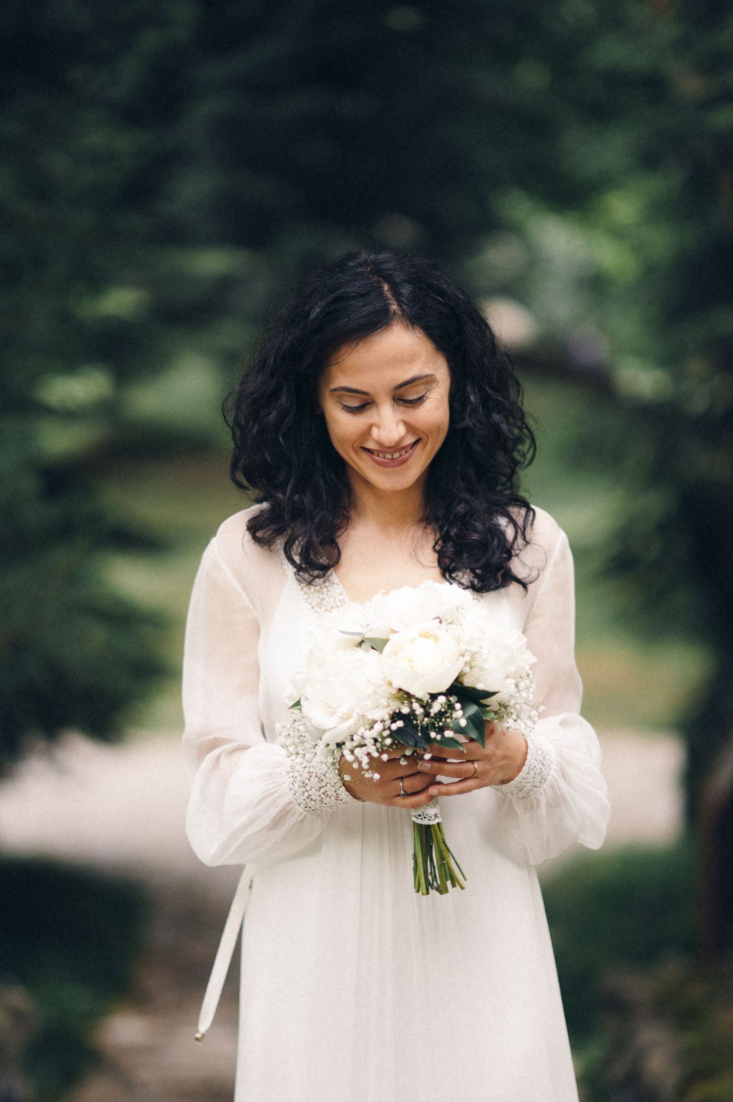 Bride in a flowing white dress smiles while holding a bouquet of white flowers in a garden setting.