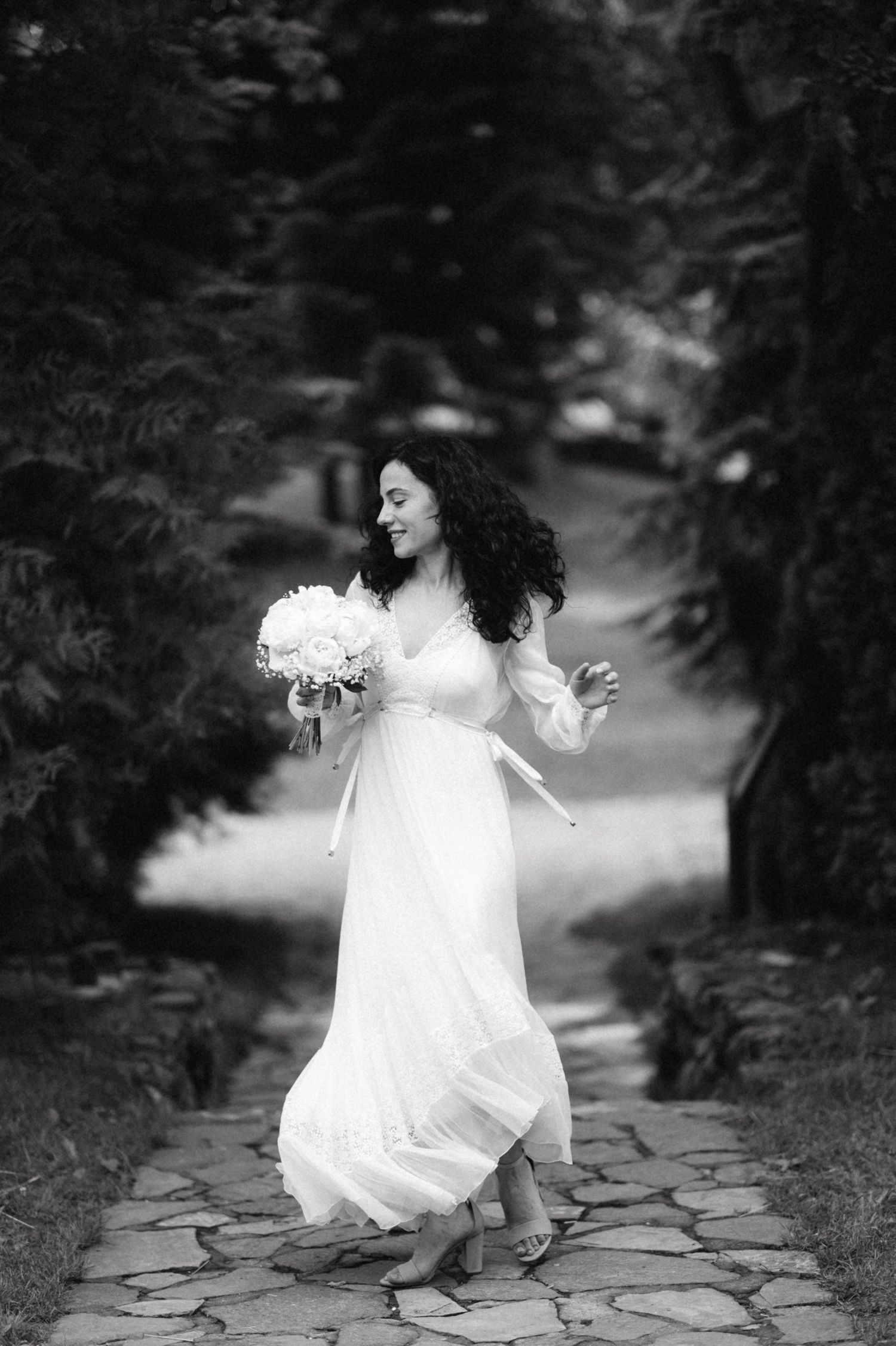 Black and white photo of a bride dancing barefoot on a stone path while holding her bouquet.